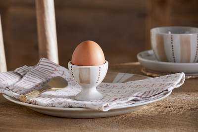 A brown egg sits upright in a nkuku Patthar Egg Cup (Cream & Clay, Set of 2), on a patterned napkin and ceramic plate. A gold spoon and matching cup and saucer complete the warm, rustic look on a wooden table.