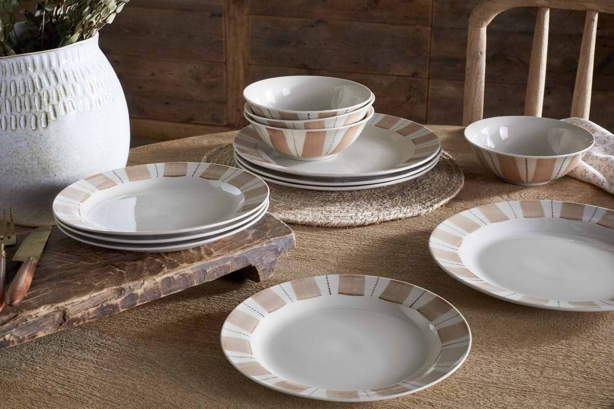 A rustic dining table set with nkuku's Patthar Dinnerware Set in Cream & Clay, featuring 12 hand-painted ceramic plates and bowls. A textured white vase with dried foliage and wooden cutlery complete the cozy scene.