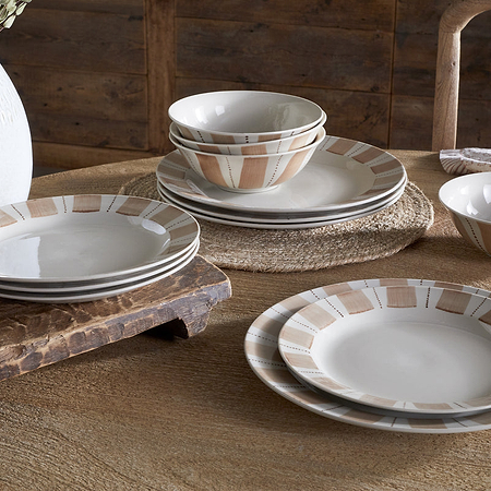 A rustic wooden table is set with nkuku’s Patthar Dinnerware Set - Cream & Clay (set of 12), some dishes stacked, others arranged. A textured white vase with dried flowers and a wooden cutting board add to the cozy dining scene.