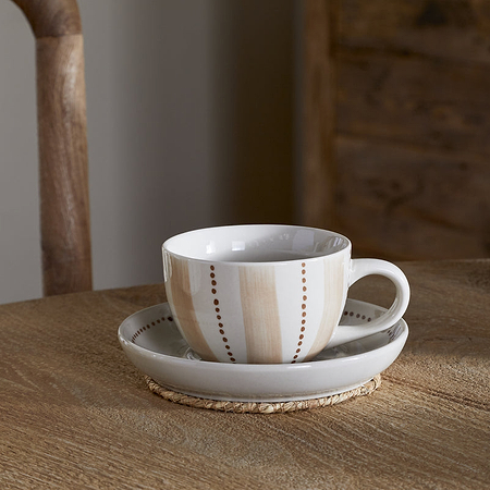 A nkuku Patthar Cup & Saucer - Cream & Clay with beige stripes rests on a wicker coaster, complemented by a matching teapot, glass jar of brown sugar, and a wooden chair and cabinet in the background.