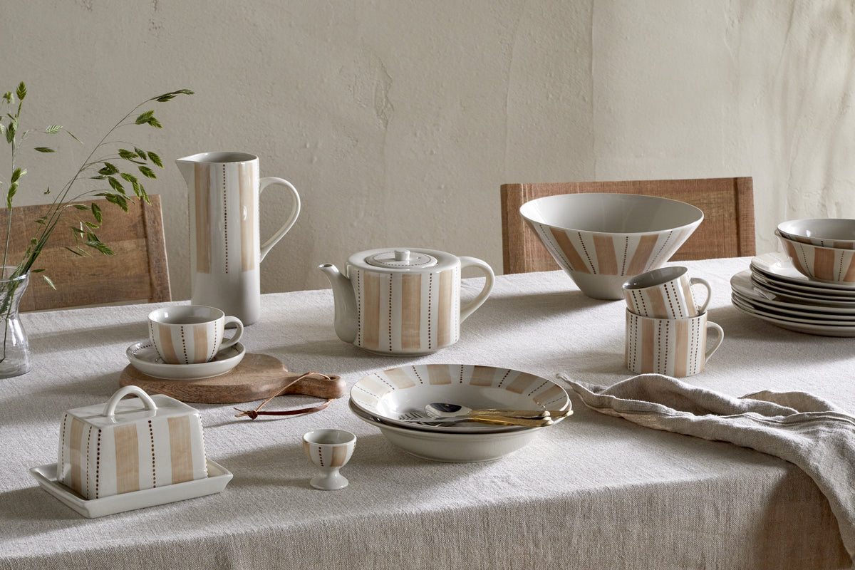A softly lit table decorated with a beige linen tablecloth, stacked nkuku Patthar Pasta Bowls in cream & clay, cream-striped ceramic dishes, cups, saucers, a teapot, pitcher, egg cup, butter dish, and a small green plant.