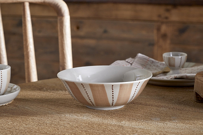 The Patthar Cereal Bowl - Cream & Clay by nkuku, featuring hand-painted tan and brown stripes with dotted accents, is displayed on a wooden table alongside coordinating pieces from the collection, a beige napkin with red stripes, and a rustic chair.