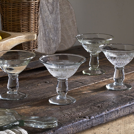 A set of four nkuku Sooraj Champagne Coupes (Clear) sits on a rustic wooden table, surrounded by lime wedges, a small bowl of salt, and fresh limes and lemons in the background for a vintage-inspired display.