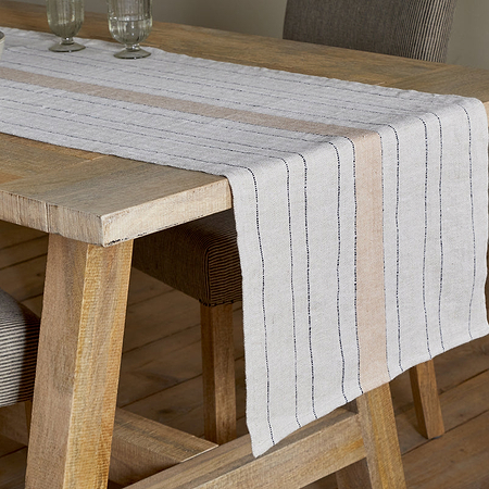 A rustic wooden dining table features the Buna Table Runner - Clay by nkuku with black and tan stripes, two glass goblets, and a large light-gray ceramic bowl, paired with striped chairs on a light wood floor.