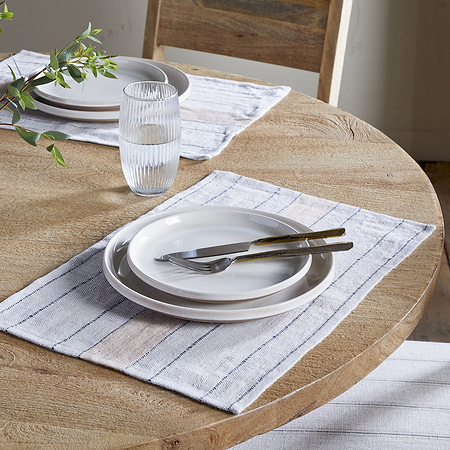 A round wooden table set for two features nkuku Buna Placemats in Clay, stacked white plates, cutlery, water glasses, a small vase with green leafy branches, and is surrounded by upholstered wooden chairs.