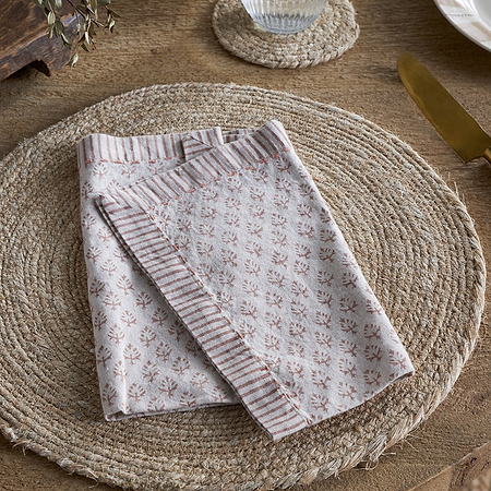 A beige woven placemat sits on a wooden table with an nkuku Peplu Block Print Napkin - Terracotta folded at the center. A gold fork is on the left, a gold knife with a wooden handle on the right, and part of a white plate is visible in the top right corner.