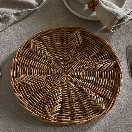 The nkuku Bersama Rattan Placemat - Natural sits on a beige linen tablecloth, with cutlery on each side. A beige napkin in a rattan ring tops stacked plates, creating a rustic dining setting.