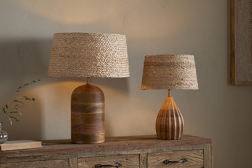 Two nkuku table lamps with Pisang Natural Lampshades sit on a rustic dresser, adding organic texture and warm light. The taller has a rounded dark brown base, while the shorter features a lighter, teardrop-shaped wooden base.