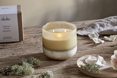 A hand-blown cream and beige glass vessel holds the glowing nkuku Oakmoss & Rose 3 Wick Scented Candle - Yellow, set on a rustic wooden table with white rose petals, green moss, and the branded box in a softly lit, serene scene.