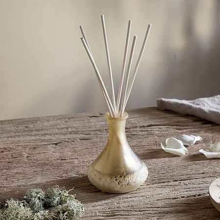 A small yellow nkuku Oakmoss & Rose Scented Reed Diffuser with white reeds sits on a rustic wooden table, surrounded by green moss, white rose petals, and its brown box, creating an inviting floral woody atmosphere.