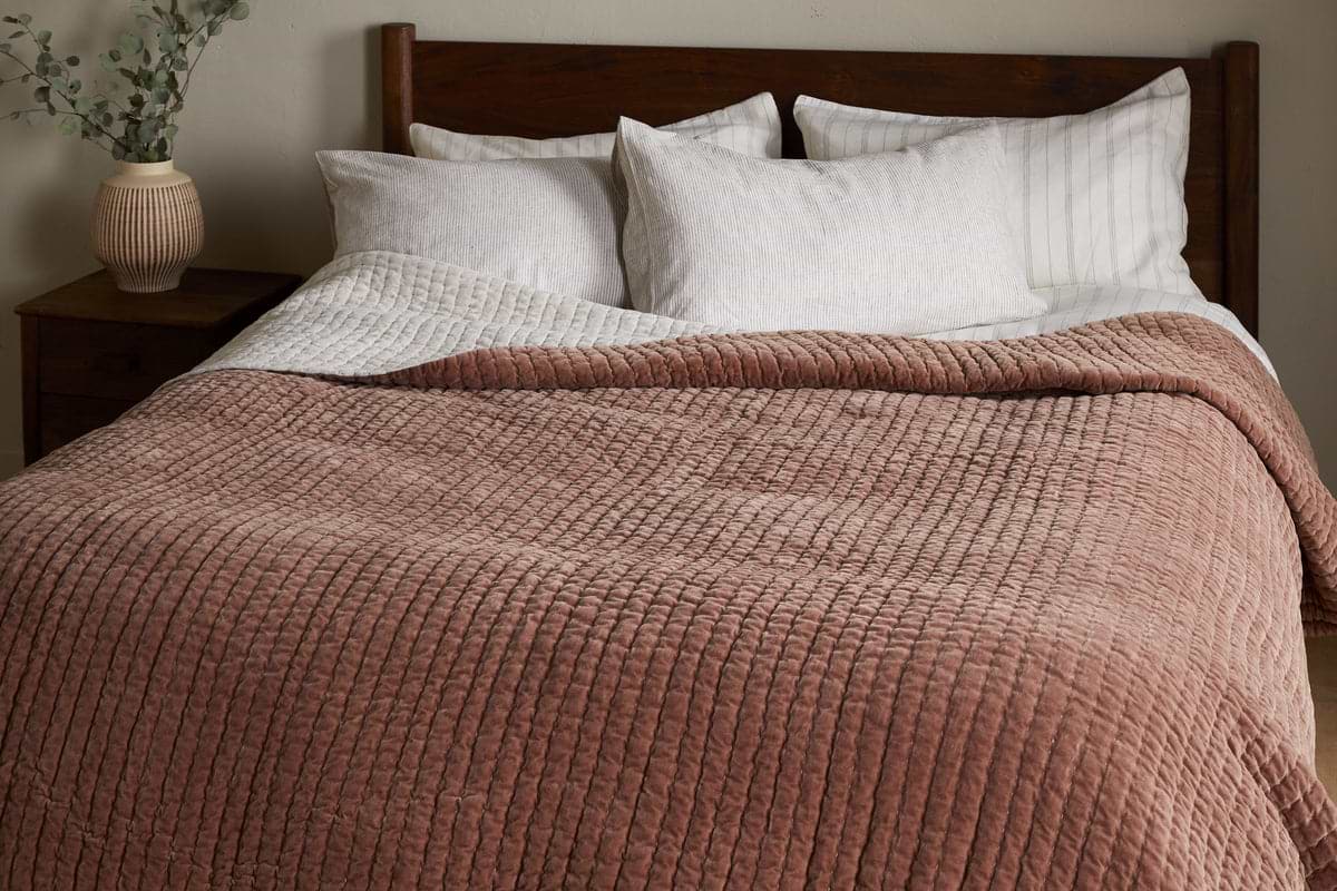 A neatly made bed with white pillows, a striped white sheet, and the nkuku Nidra Velvet & Linen Bed Quilt in Rust. A wooden headboard and a nightstand with greenery complete this King Super King bedding setup.