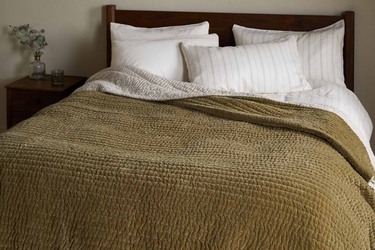 A neatly made bed with white pillows and linens is partially covered by the nkuku Nidra Velvet & Linen Bed Quilt in Moss. Against a wooden headboard, a small nightstand holds a glass vase of eucalyptus and a candle.