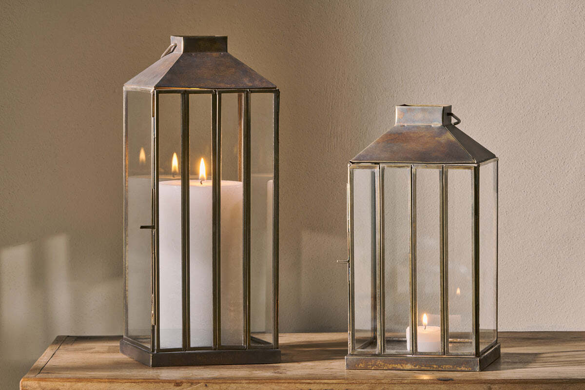 Two nkuku Nampo Lanterns - Aged Antique, one tall with a pillar candle and one short with a tealight, cast warm light from their brass and glass frames onto a wooden surface against a beige wall.