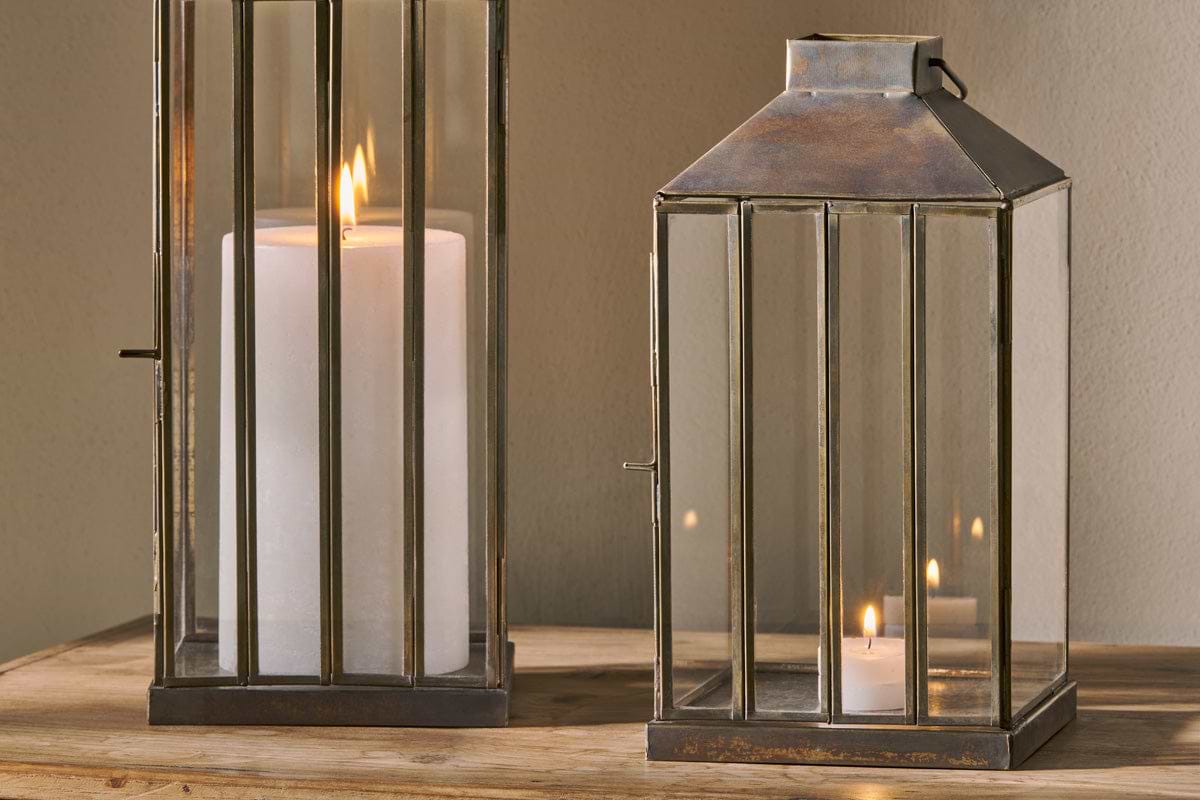 Two nkuku Nampo Lanterns - Aged Antique, made of brass and glass, sit on wood. The left contains a large white pillar candle with a tall flame; the right holds a smaller lit candle, both casting a warm glow against a neutral background.