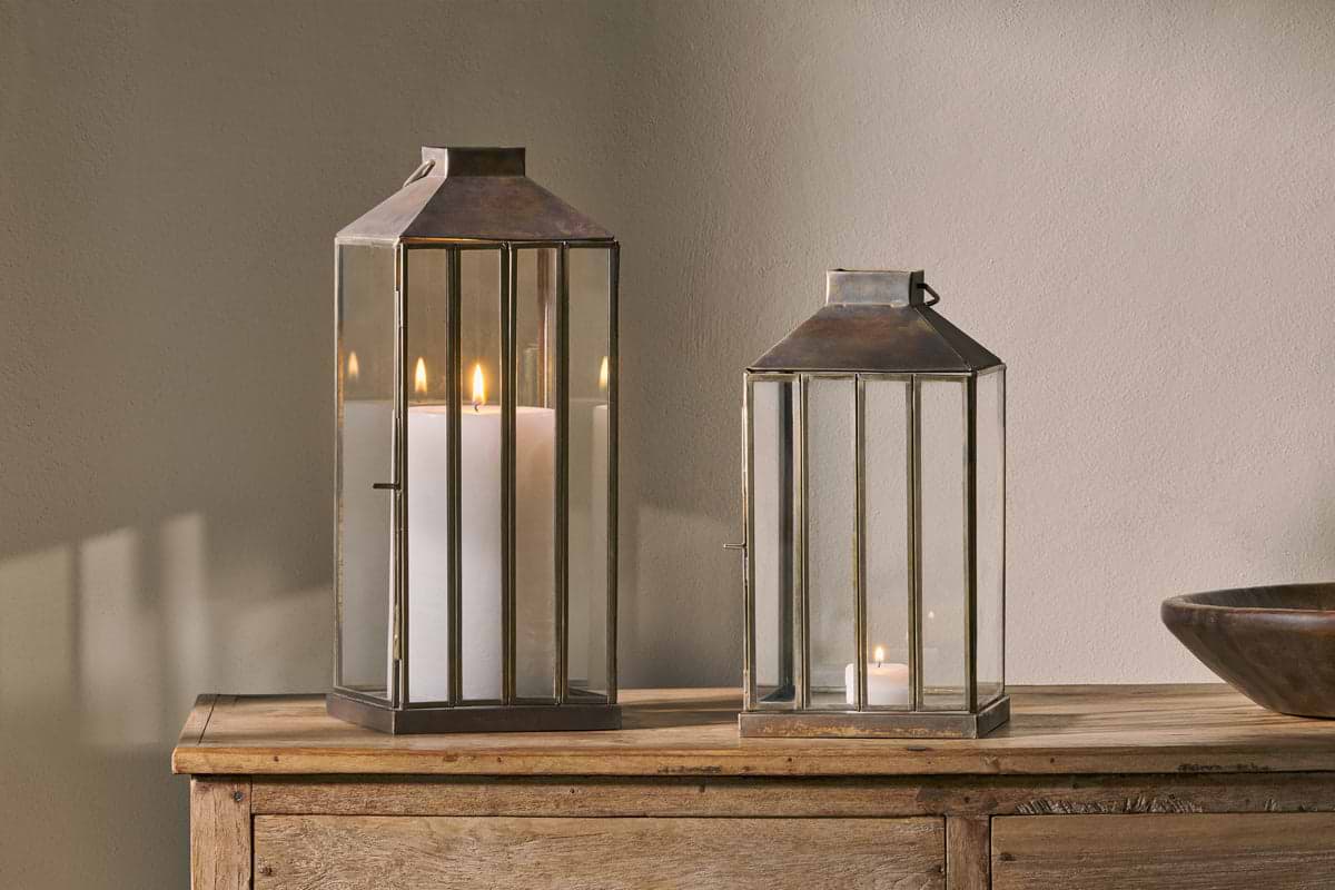 Two nkuku Nampo Lanterns - Aged Antique, one tall with a pillar candle and one short with a tealight, sit on a wooden surface. Warm candlelight glows through the brass and glass, casting soft shadows on the wall behind them.