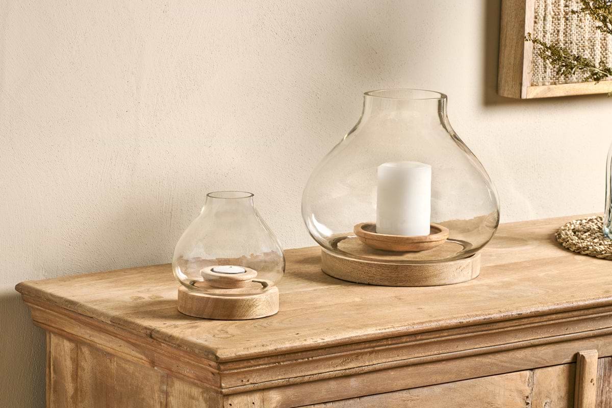 Two nkuku Naryla Mango Wood Lanterns made of recycled glass and natural mango wood sit on a rustic cabinet—one with a pillar candle, one with a tealight. A framed textured wall art is partially visible in the upper right corner.