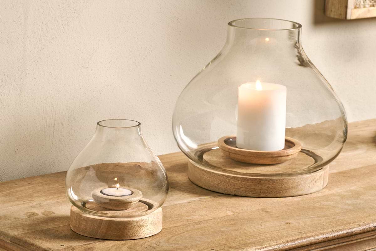 Two nkuku Naryla Mango Wood Lanterns in natural finish, one with a tealight and the other with a pillar candle, cast warm light from their mango wood bases and recycled glass tops on a wooden surface against a plain wall.
