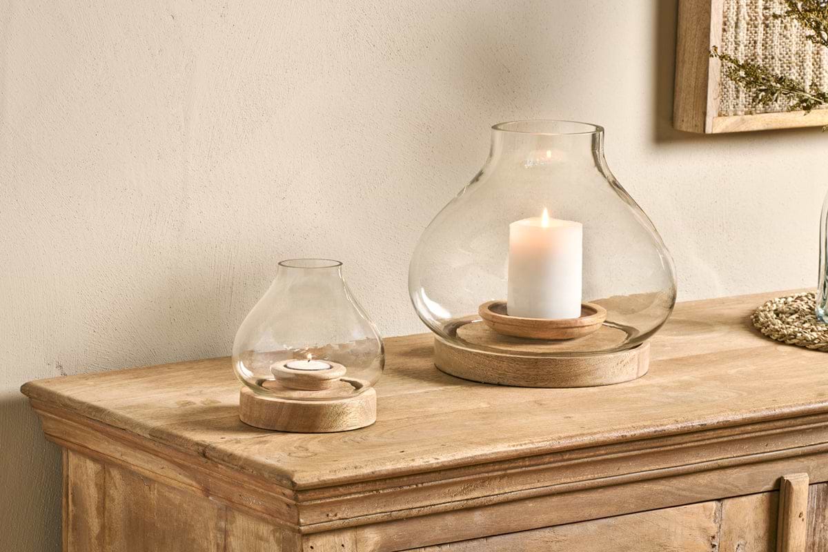Two nkuku Naryla Mango Wood Lanterns in natural, each with a recycled glass top and mango wood base, sit on a rustic cabinet; the larger holds a pillar candle, the smaller cradles a tealight against a neutral-toned wall.