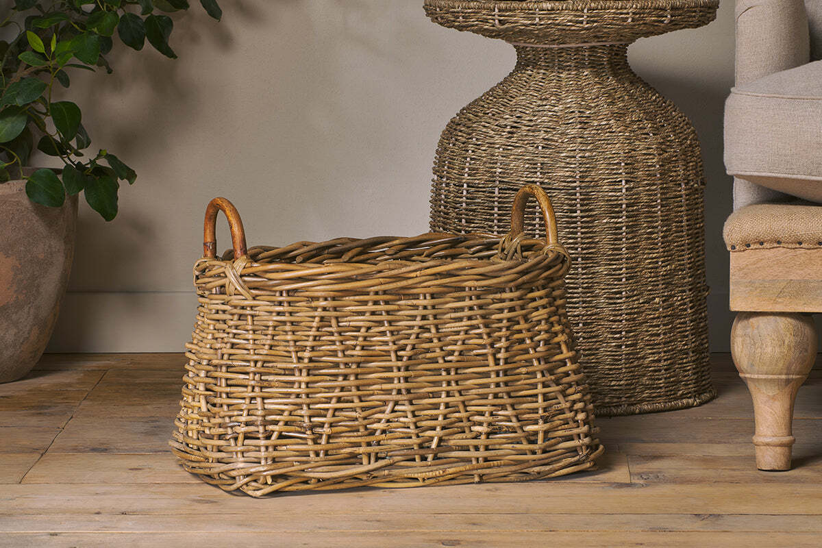 The Netra Rattan Basket - Natural by nkuku, a large rectangular basket with two looped handles, adds rustic charm as decorative storage in your home—perfect beside a beige sofa and potted plant for cozy style.