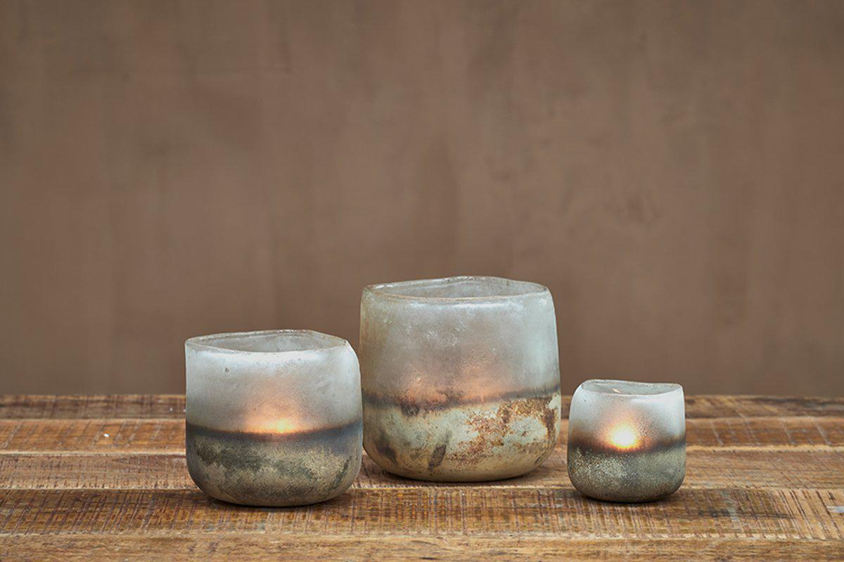Three uneven Ngolo Tealight Holders in aged smoke from nkuku, crafted from frosted recycled glass, glow with lit candles on a rustic wood surface. The plain brown wall behind enhances their cozy, minimalist atmosphere.