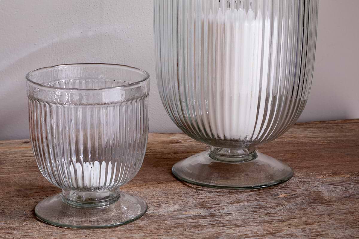 Two Nia Recycled Glass Candle Holders by nkuku, featuring a fluted design and flared bases, sit on a rustic wooden surface. The taller clear holder contains white powder; the smaller is empty. Both can also be used as unique goblets.