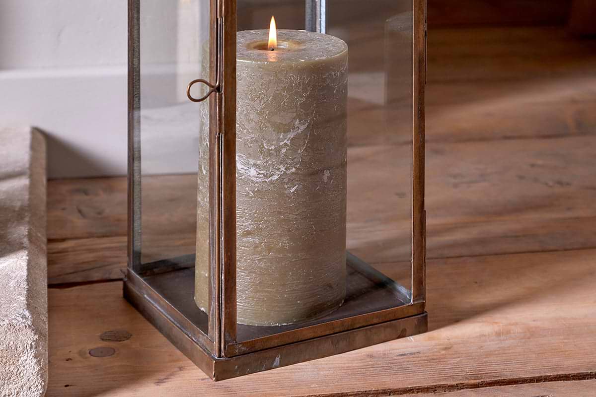 The Nivas Lantern - Antique Brass by nkuku sits on a wooden floor, glowing with a steady pillar candle. Warm natural light highlights this elegant Eastern-style home accessory.