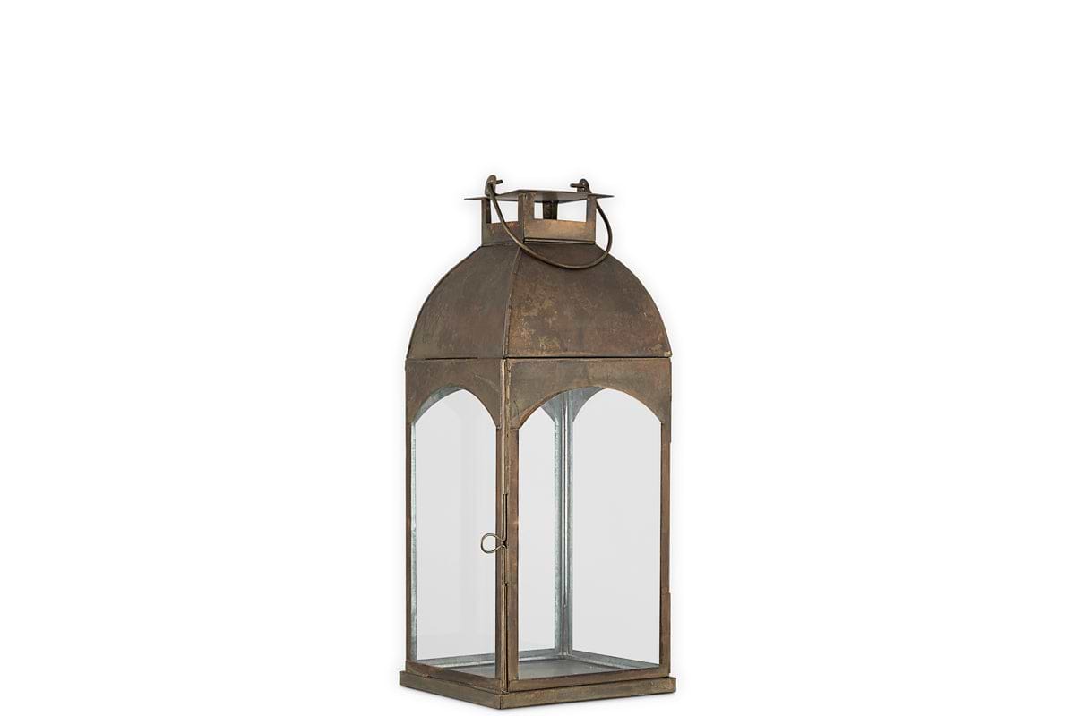 The Nivas Lantern by nkuku features an antique brass finish, arched glass panels on all sides, and a round handle on top—a vintage-style home accessory that adds timeless charm to any space.