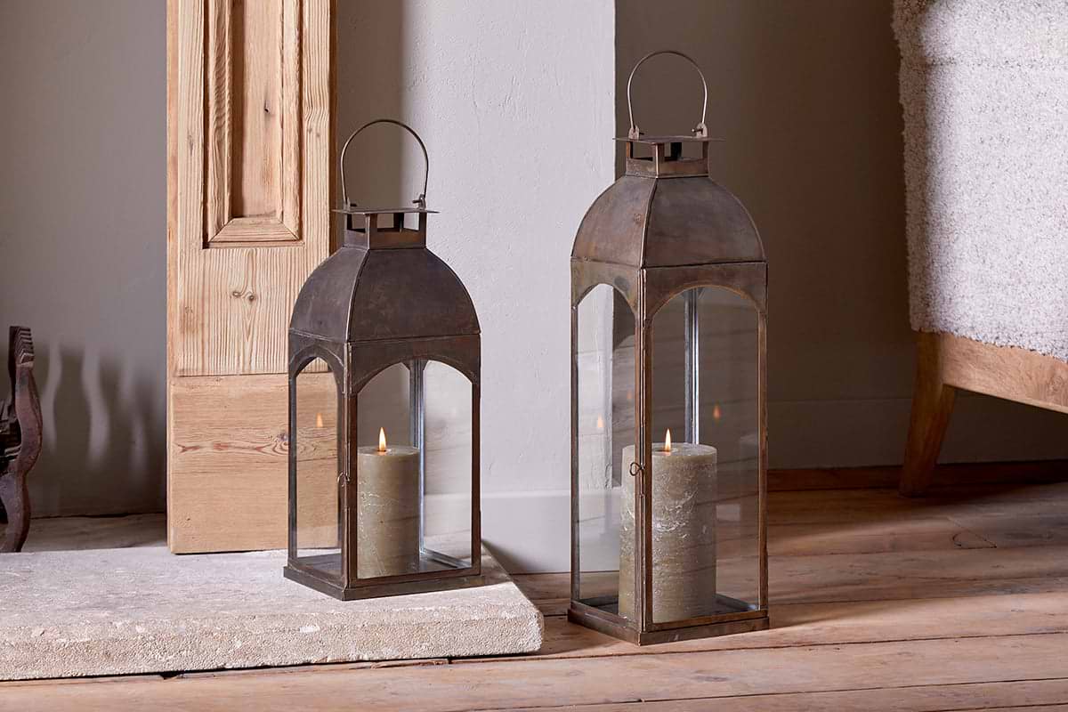 Two Nivas Lanterns in Antique Brass by nkuku, each with a glowing pillar candle, sit on a wooden floor beside a stone hearth and wood paneling. Neutral walls and part of a textured beige chair complete the cozy scene.