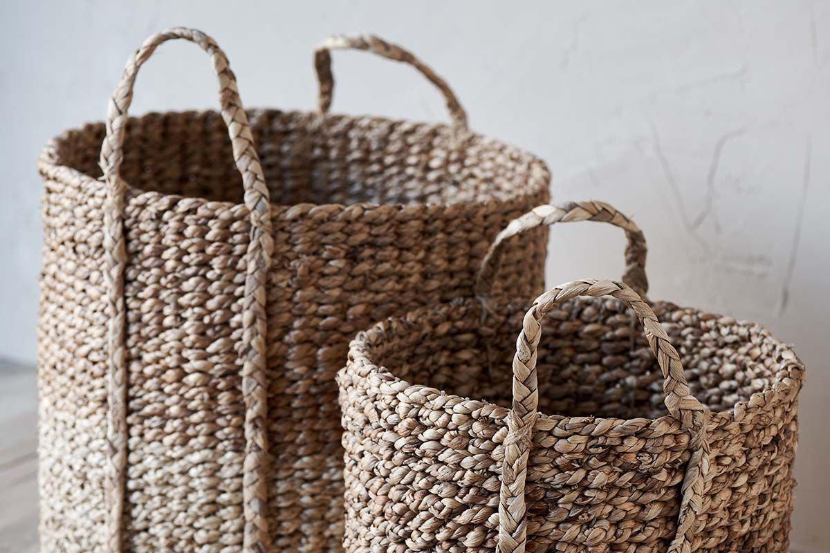 Two nkuku Nkomi Baskets, hand woven from natural textured fibers, sit side by side on a light floor. Both cylindrical storage baskets feature sturdy handles; the larger one is behind the smaller, set against a pale wall.