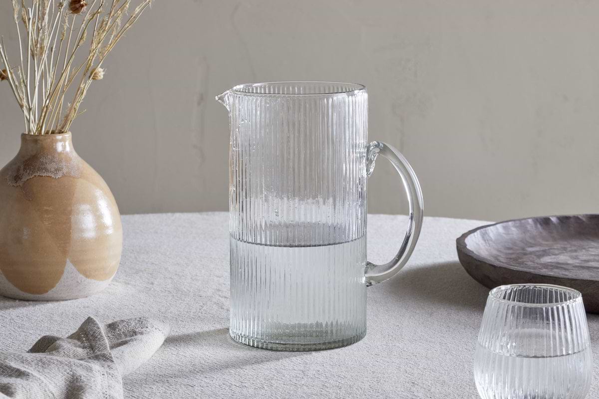 An Otuba Jug - Clear by nkuku, half-filled with water, rests on a light-colored textured tablecloth beside a matching glass, a beige ceramic vase with dried flowers, a napkin, and a wooden bowl.