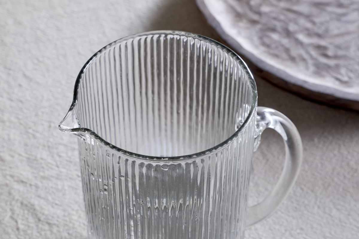 The Otuba Jug - Clear by nkuku, made from recycled glass with a vertical ribbed texture, rests elegantly on a light surface. In the background, a subtly patterned white plate complements its timeless design.