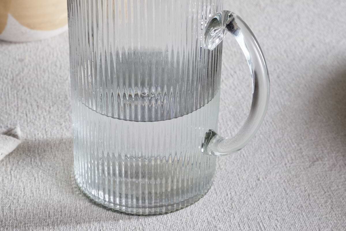 A close-up of the Otuba Jug - Clear by nkuku, featuring a ribbed design and crafted from recycled glass, half-filled with water and set on a textured light-colored tablecloth.