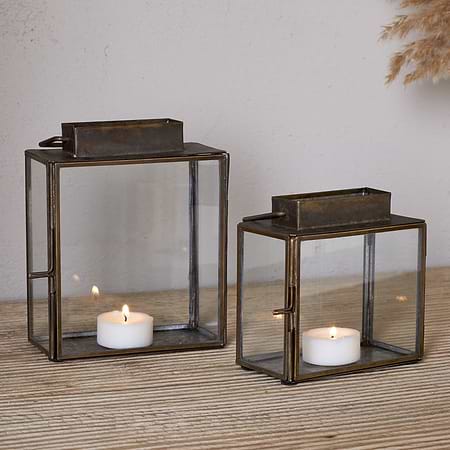 Two nkuku Obi Mini Table Lanterns in antique brass, each with a metal frame and glass door holding a lit tealight, sit on a wooden surface. A vase of dried pampas grass in the background adds to the candlelit ambiance.