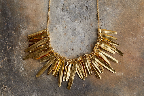 The nkuku Omuti Necklace - Gold features multiple elongated, flat, textured pendants in a bold gold finish, hanging closely together on a chain and displayed on a stone surface.