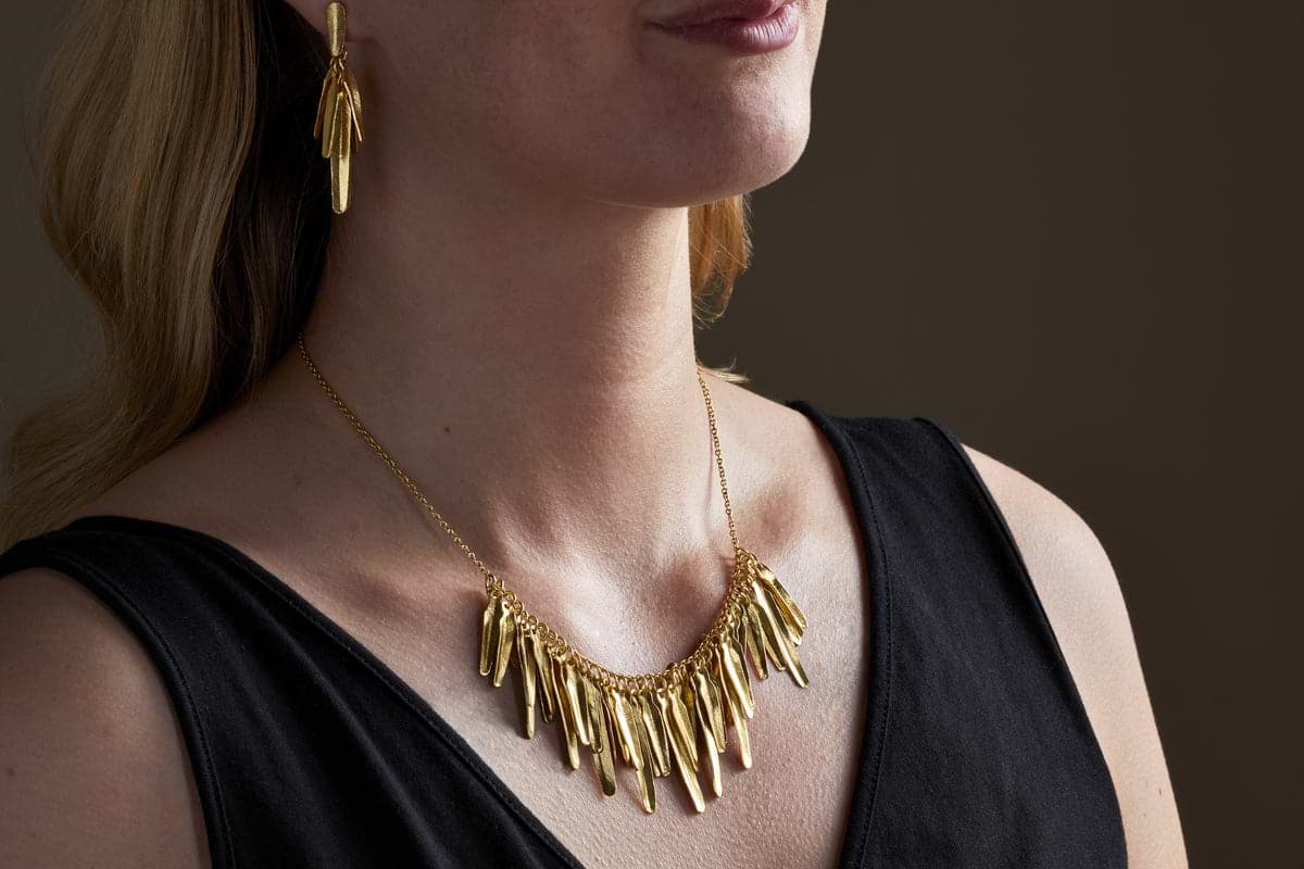 A woman in a black sleeveless top models the nkuku Omuti Necklace - Gold, paired with matching gold earrings featuring bold, elongated shapes, set against a dark background.