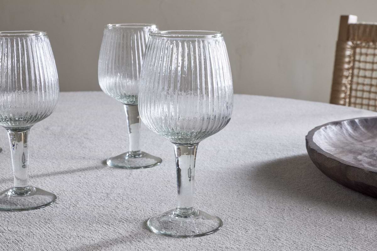 Four elegant, ribbed Otuba Wine Glasses by nkuku sit on a light textured tablecloth, with a wooden bowl and woven chair in the background for a serene, minimal look.