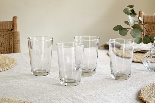 The nkuku Pani Highball - Clear (Set of 4) glassware is displayed on a light tablecloth, paired with woven placemats, wooden chairs with woven seats, and a vase of eucalyptus for a relaxed dining vibe.
