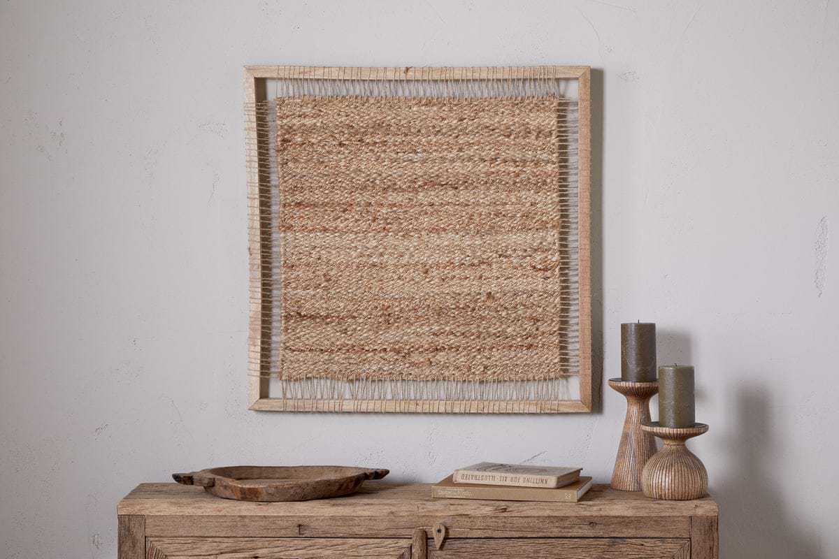 The nkuku Pemali Woven Jute Wall Art - Natural is displayed above a rustic wooden sideboard with wooden candlesticks holding gray candles, a shallow tray, and a book for cozy decor against a white wall.