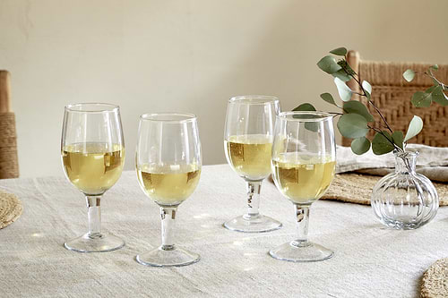 The nkuku Pani Wine Glass - Clear (Set of 4) sits on a light tablecloth with white wine. A vase with eucalyptus branches is nearby, and woven chairs in the background create a perfect everyday dining setting.