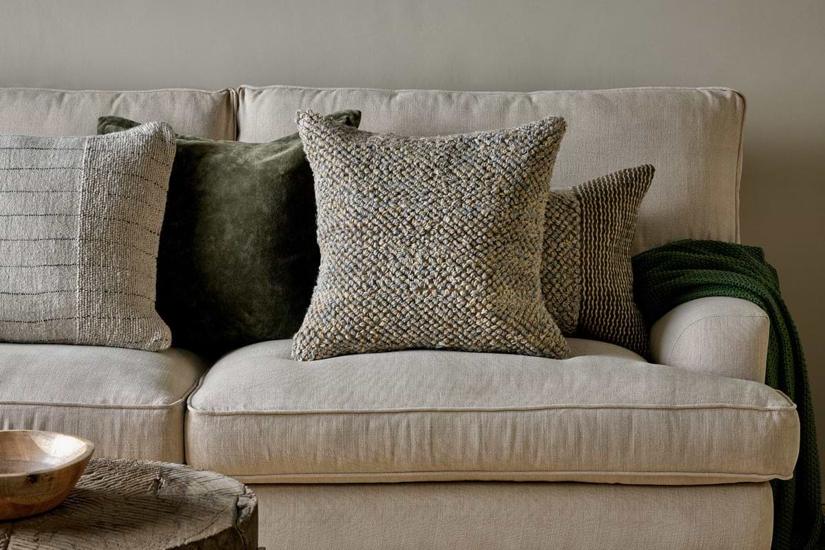 A beige sofa with feather cushion inners is styled with four decorative pillows in soft gray, green, and the nkuku Paduna Jute & Cotton Cushion Cover in soft blue & brown. A dark green throw drapes the right arm; a wooden bowl rests on a rustic coffee table.