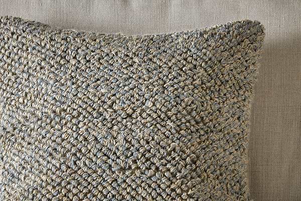Close-up of the nkuku Paduna Jute & Cotton Cushion Cover in soft blue and brown, featuring a chunky woven texture. It rests on a taupe fabric background, its tactile look complemented by plush feather inners for added comfort.