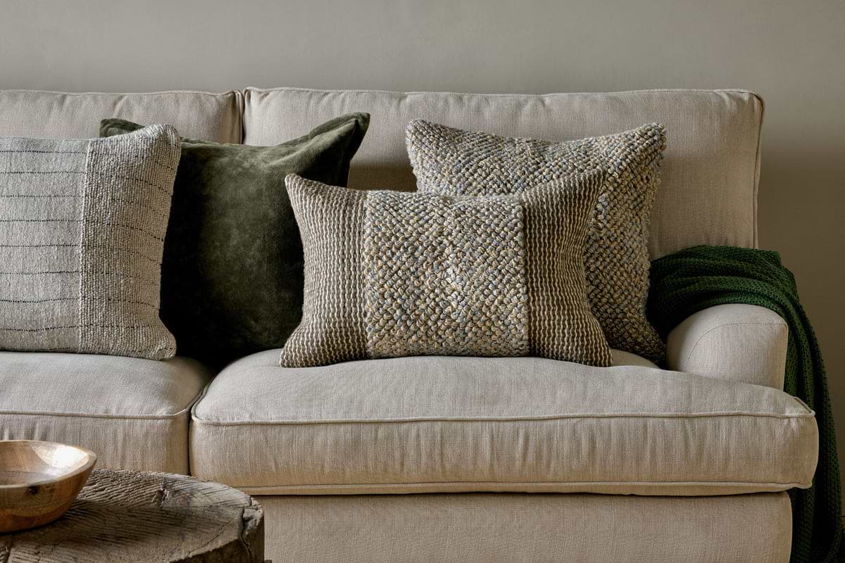A beige sofa displays four nkuku Paduna Jute & Cotton Cushion Covers in soft blue and brown, each with feather inners. A green knit blanket drapes over the armrest, and a rustic wooden coffee table holds a light wooden bowl.