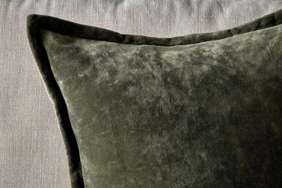 A close-up of the nkuku Pilani Velvet & Linen Cushion Cover in Moss, showcasing its soft, plush velvet texture and piped edges as it rests on a light gray fabric surface with lighting highlighting its subtle sheen.