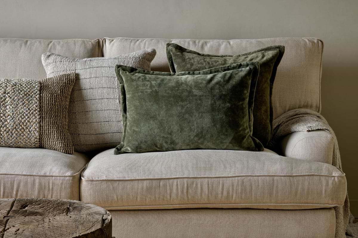 A beige linen sofa features textured throw pillows in neutral and olive tones, including nkuku’s Pilani Velvet & Linen Cushion Cover in Moss. A knit blanket rests on the arm beside a rustic wooden coffee table, set against a light gray wall.