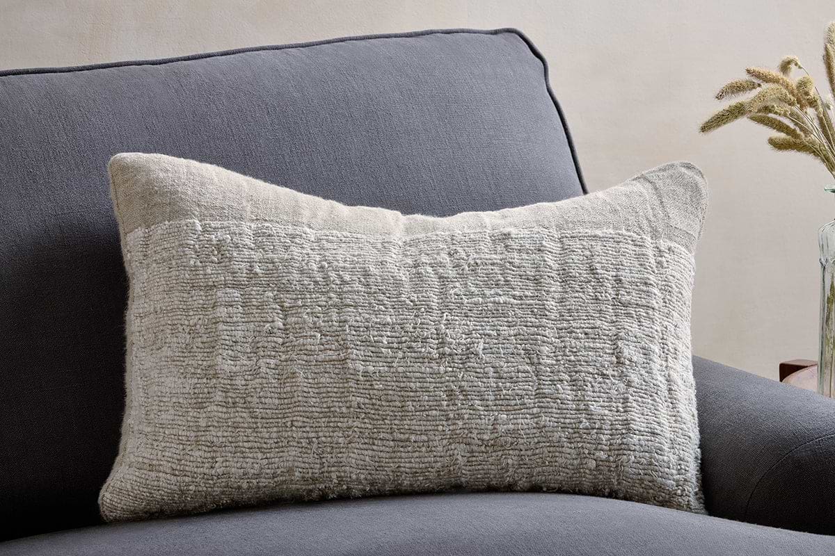 The nkuku Puli Recycled Linen Cushion Cover in Natural, with its soft textured beige linen, rests on a dark gray armchair. Light walls and a glass vase with dried grasses are partly visible in the background.