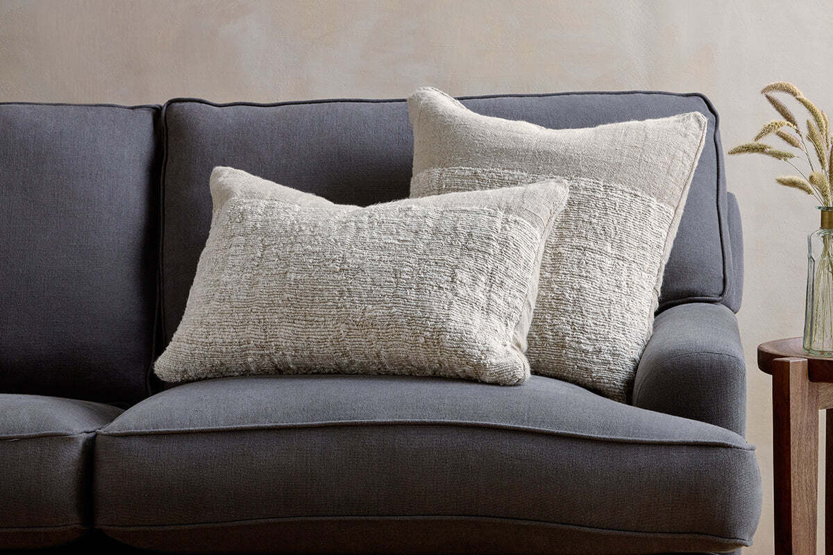 A dark gray upholstered couch features two nkuku Puli Recycled Linen Cushion Covers in natural, arranged against the backrest. On the right, a small wooden side table holds a glass vase with dried grasses. The wall behind is light and neutral.