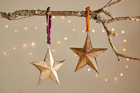 The nkuku Roshani Star Baubles (Set of 2) in antique brass hang from a lichen-draped branch with purple and orange ribbons, while warm fairy lights softly glow in the background, creating a cozy festive ambiance.