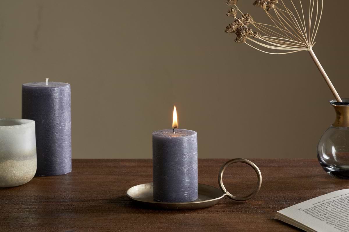 A lit nkuku Rustic Soy Blend Mini Pillar Candle in charcoal, with a textured finish, sits on a bronze handled holder atop a wooden table. Nearby are an unlit candle, a ceramic cup, a dried flower vase, and an open book.