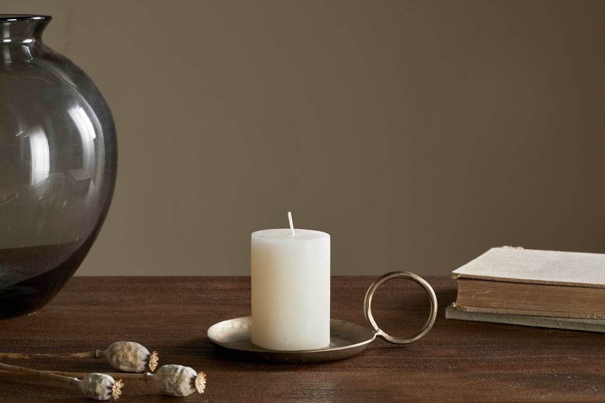 The nkuku Rustic Soy Blend Mini Pillar Candle in white, featuring a textured finish, rests on a brass holder atop a wooden table beside a closed book, dried poppy pods, and a dark glass vase against a beige background.
