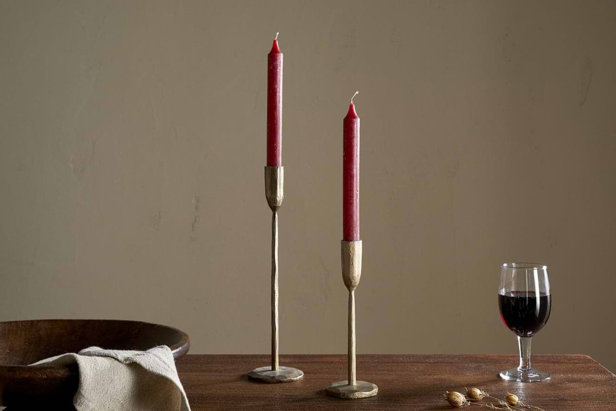 A set of two nkuku Rustic Soy Blend Dinner Candles in Dark Cherry stands on a wooden table beside a glass of red wine, beige napkin, wooden bowl, and dried berries against a plain beige wall.